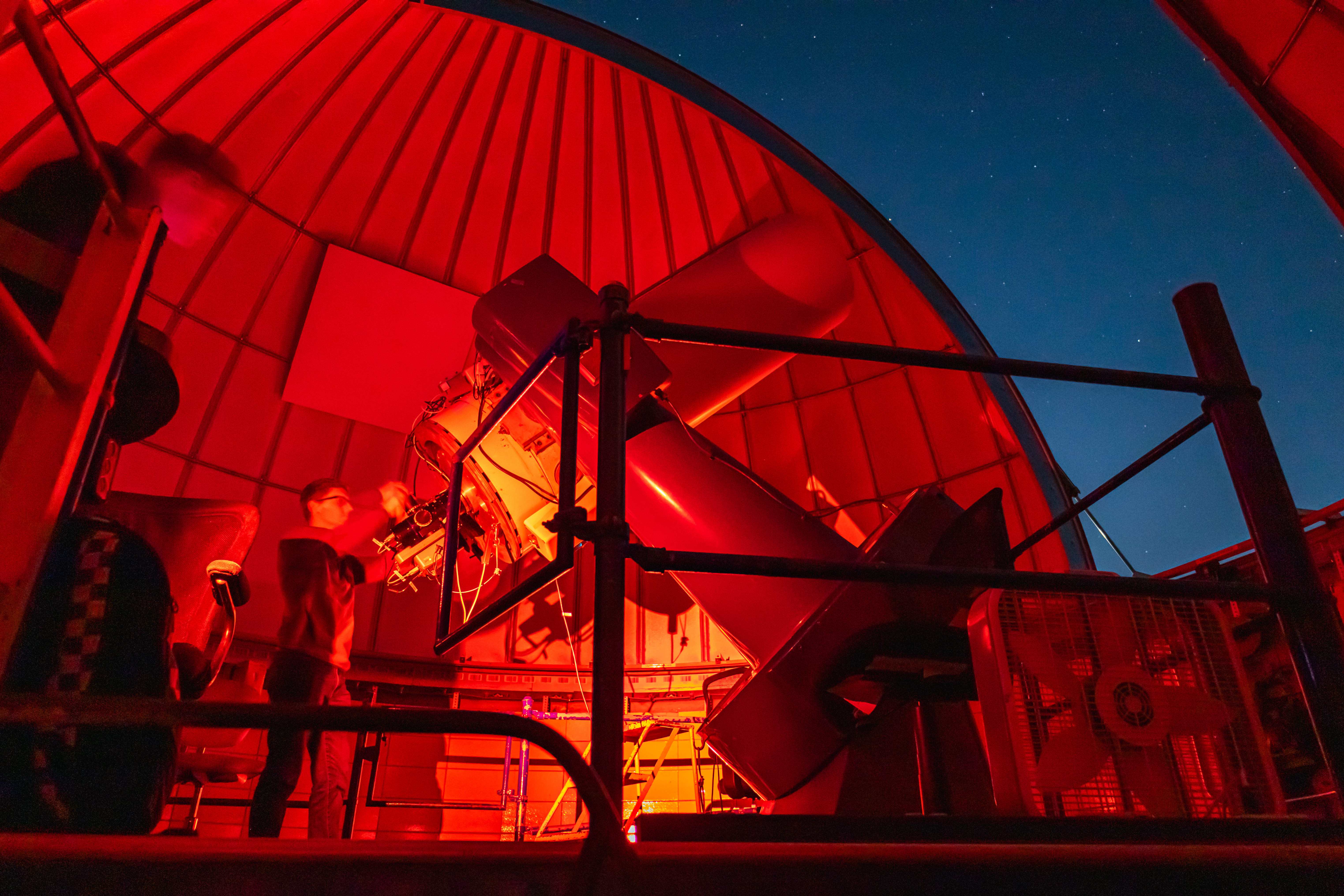 Inside the 24 inch dome lit with red light with the dome slit open to a dark blue sky with telescope operators. Photo by Brian Finch.