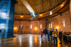 A group of visitors listen to a tour guide talk about the Great Refractor Telescope while standing in the dome.