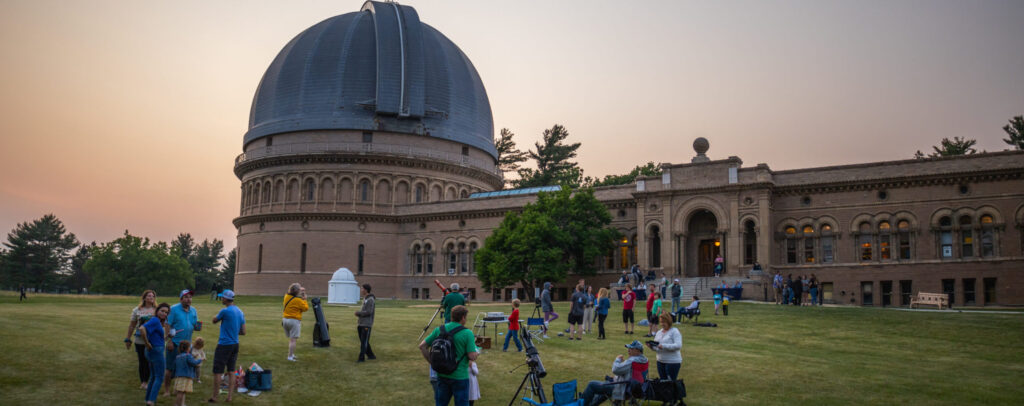 Yerkes Observatory at dusk with visitors observing through telescopes on the lawn outside.