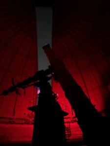 The Great Refractor telescope pointing at the night sky through the dome's shutter in red light.