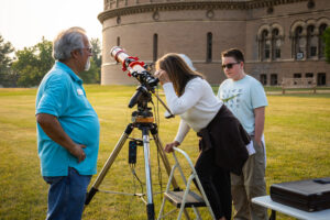 A woman looks through a telescope in front of Yerkes Observatory with a man and boy looking on.