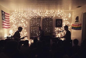 Musicians including S. Carey play in a dark room with Christmas lights and flags on the walls behind them.