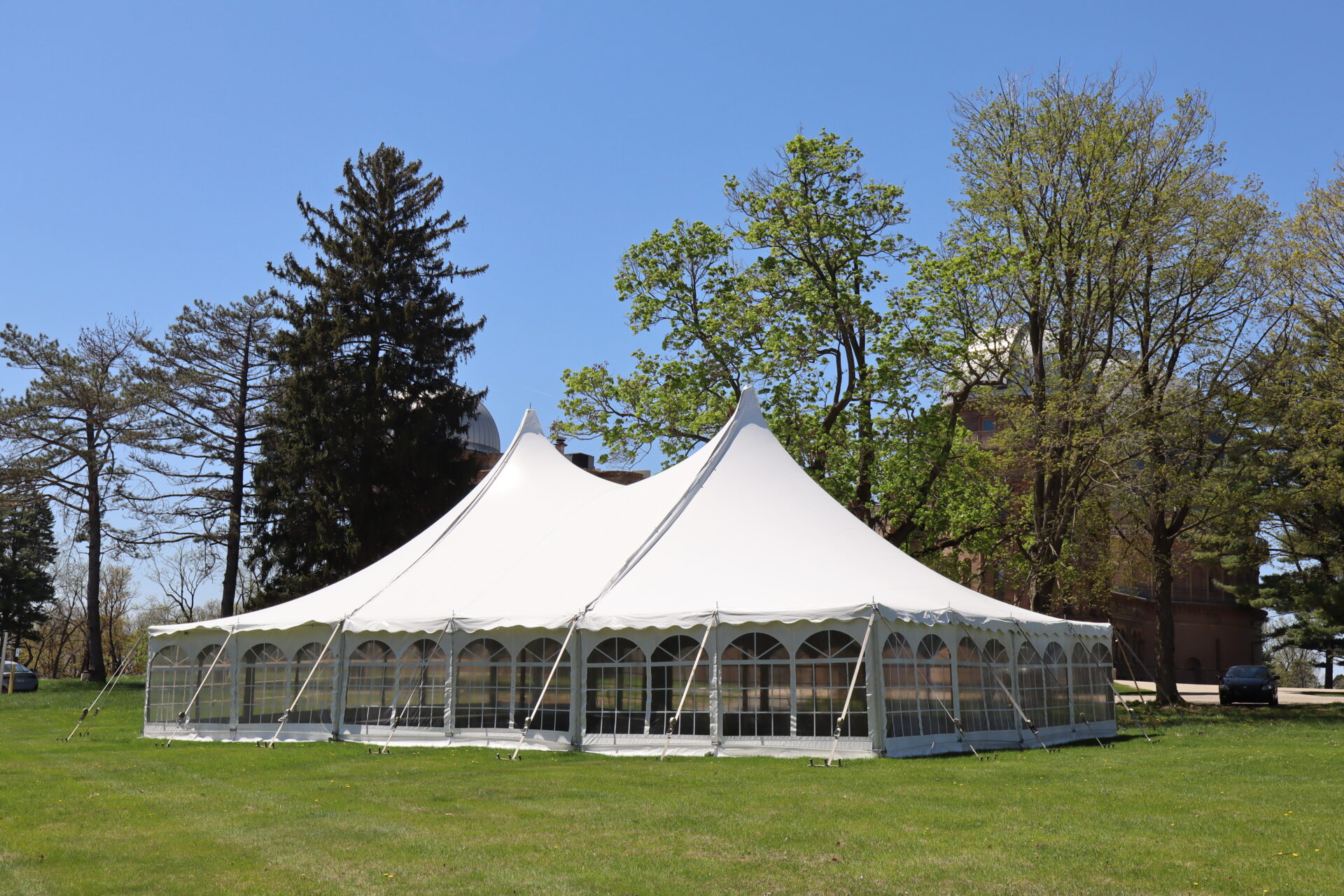 A white event tent on the grass with trees and a building in the background.