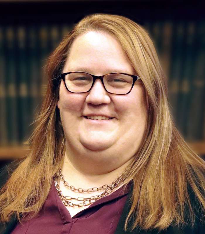A person wearing glasses with long brown hair, a maroon shirt, and several chain necklaces in front of a dark background.