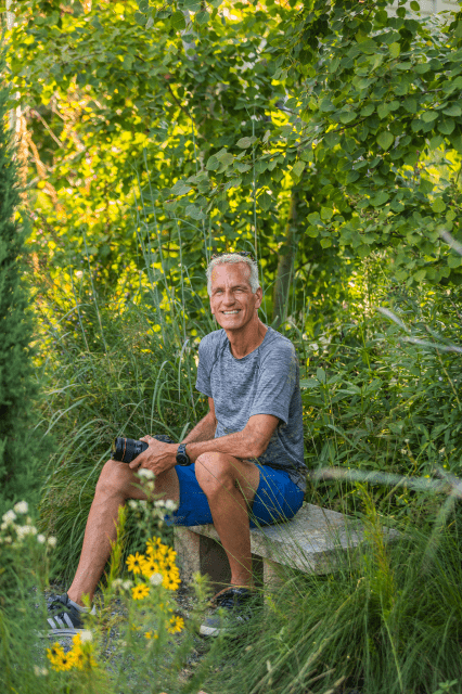 A smiling person wearing a blue t-shirt and blue shorts in a garden