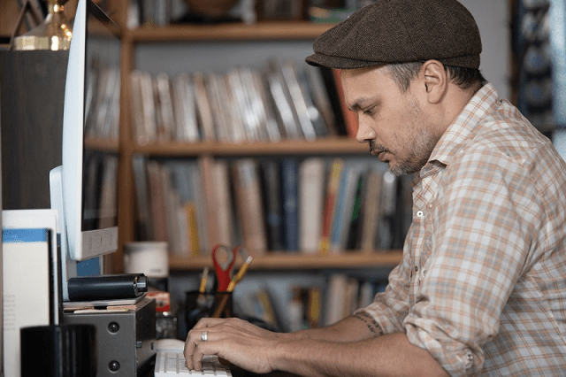 Profile view of a person wearing a brown cap and a short sleeve plaid shirt at a desk with a bookshelf in the background.