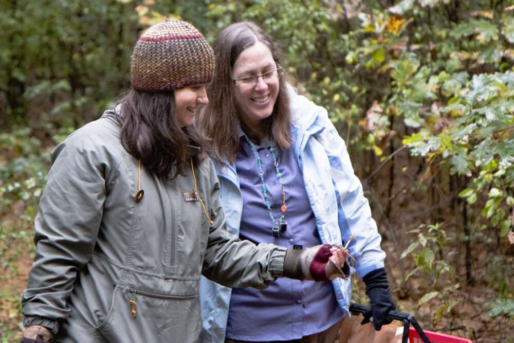 Two smiling people in the woods, looking towards the ground.